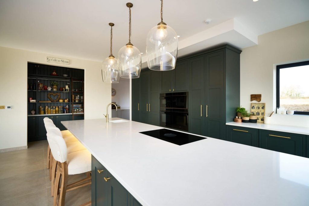 Modern kitchen with dark green shaker cabinets with brushed gold accents, a large island with a sleek white counter, featuring pendant lights and a pantry nook.