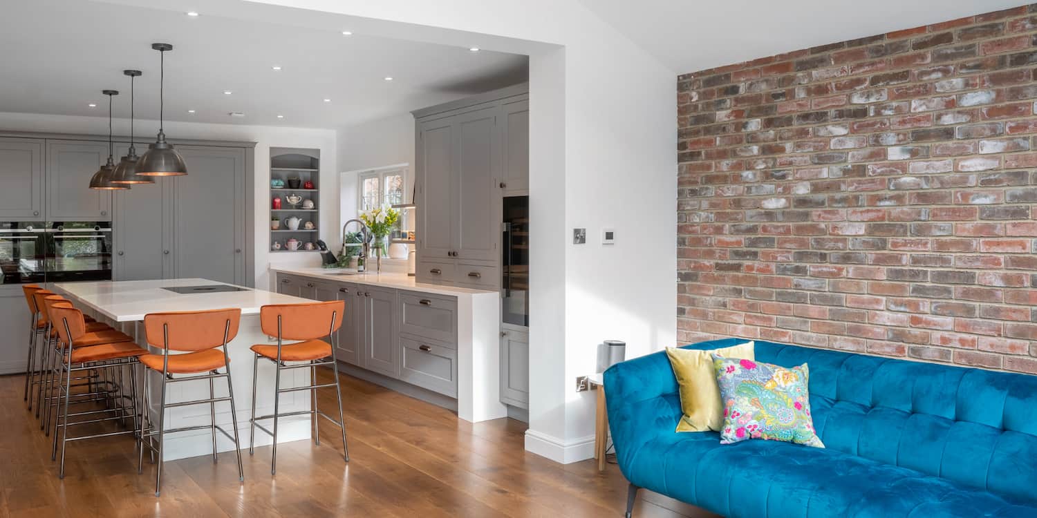 Kitchen with a large white island, grey cabinetry, orange bar stools, and a blue sofa, featuring an exposed brick wall.