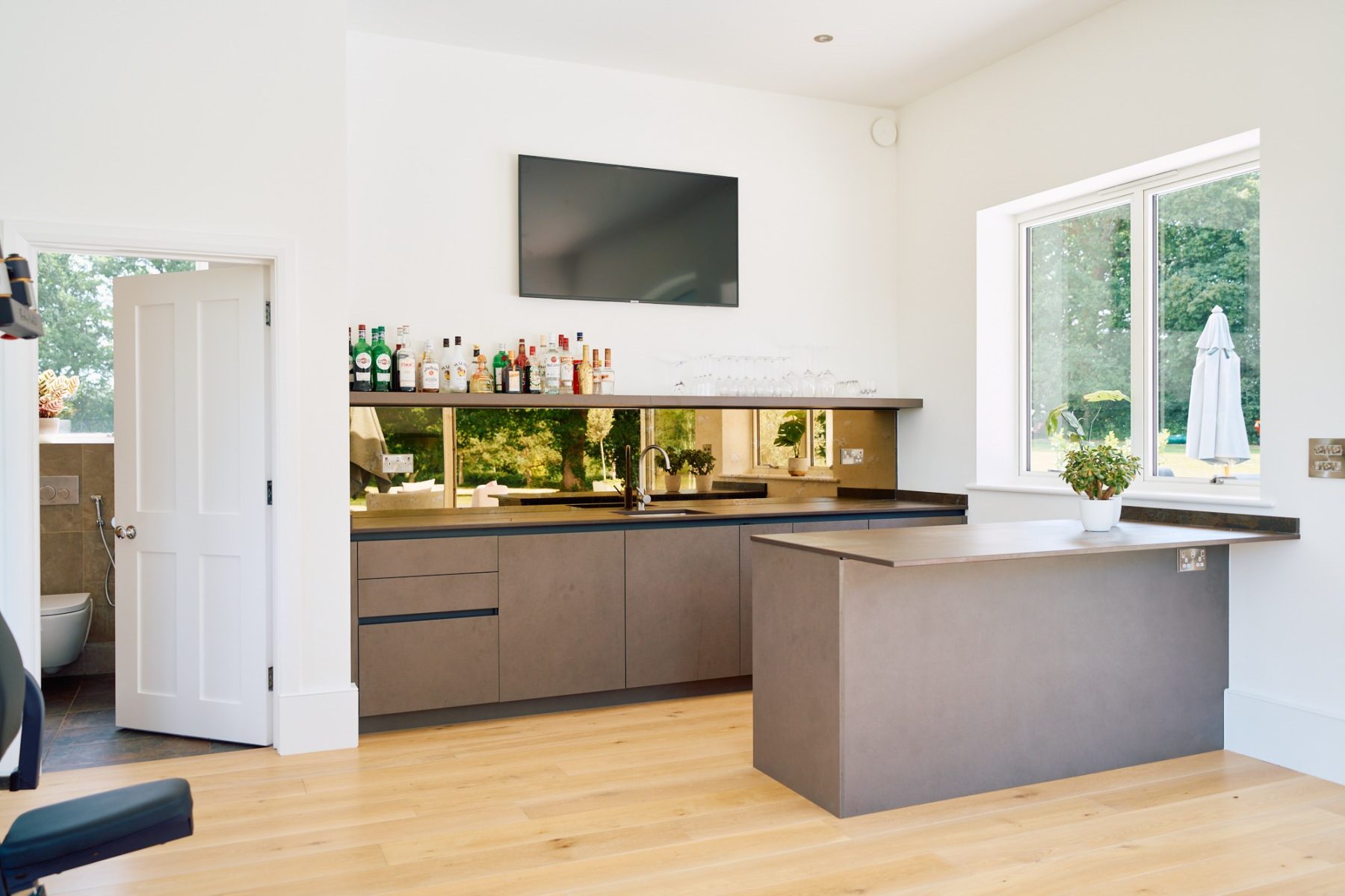 Minimalist home bar with bronze mirror splashback, matte grey cabinetry, and oak flooring in a contemporary open-plan home.