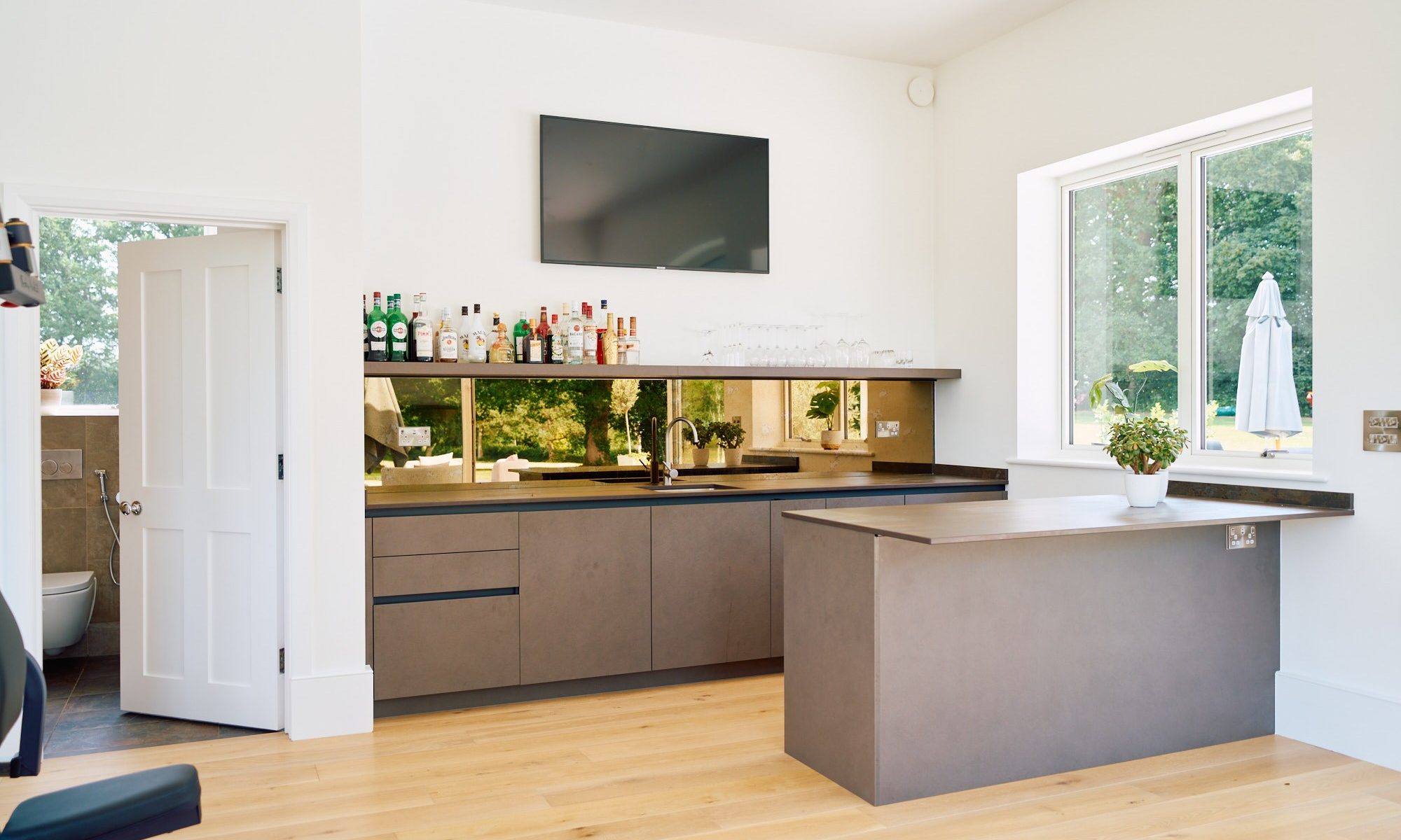 Minimalist home bar with bronze mirror splashback, matte grey cabinetry, and oak flooring in a contemporary open-plan home.