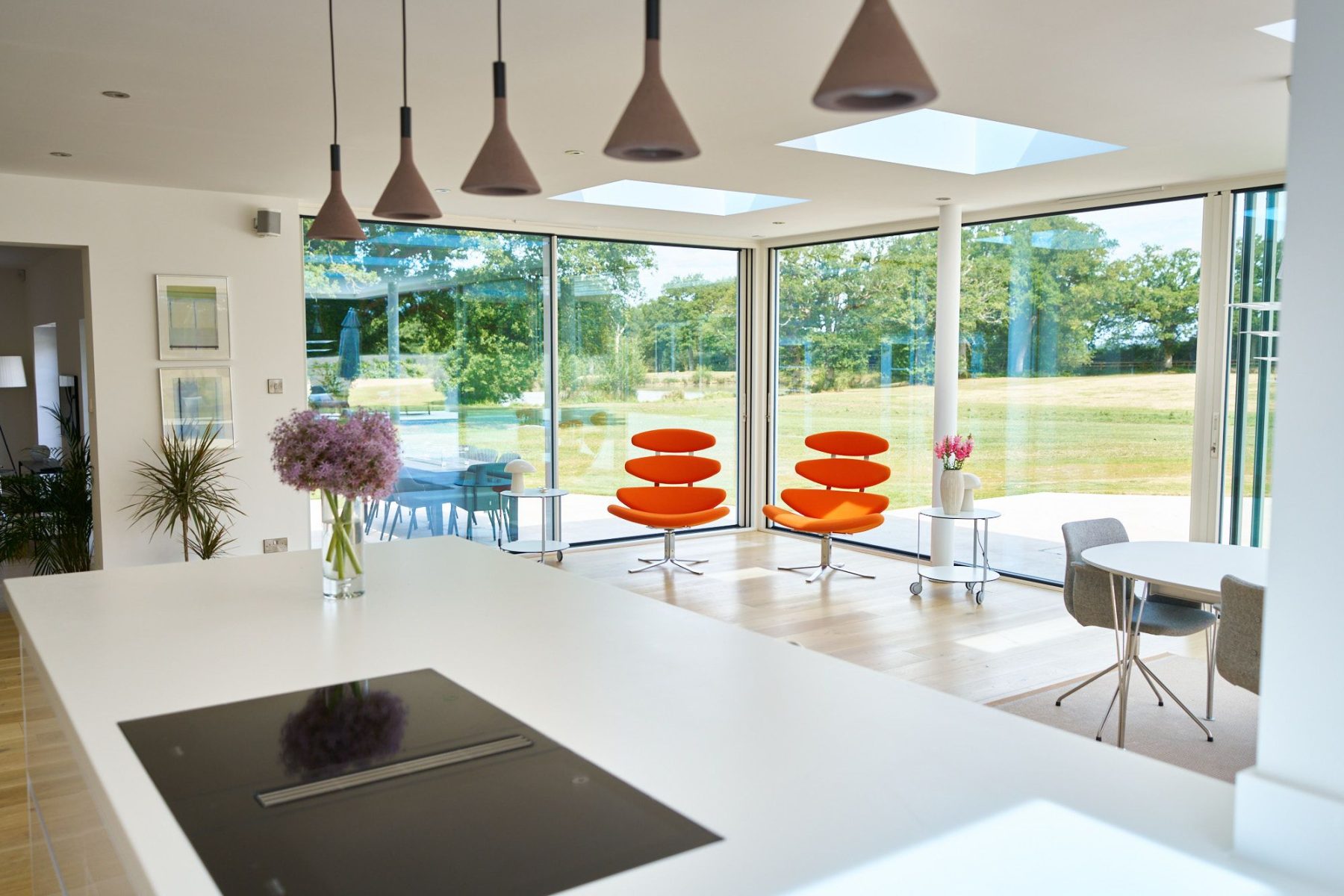 Bright open-plan white kitchen with large island, modern pendant lighting, and bold orange accent chairs overlooking garden views.
